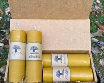 May include: A cardboard box filled with several yellow pillar candles. Each candle has a white label with a tree logo and the text "FewEssentials." The candles are cylindrical and have a clean, natural appearance. The box is open, revealing the candles inside.
