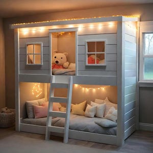 May include: A white wooden bunk bed designed like a house with a ladder, featuring two levels with mattresses, pillows, and string lights. The top level contains a plush bear toy, while the bottom level has multiple pillows and warm lighting.