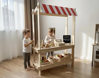 Può includere: Un banco di mercato in legno per bambini con una tenda a strisce rosse e bianche. Il banco ha un cartello di lavagna che dice "KIDS MARKET". Ci sono cesti di frutta e verdura di legno sugli scaffali. Due bambini stanno giocando con il banco.