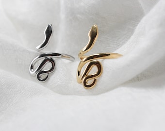 May include: Two snake-shaped rings, one silver and one gold, on a white fabric background.