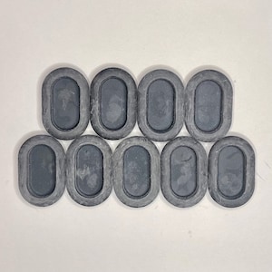 May include: A collection of ten oval-shaped, dark gray rubber grommets arranged on a white surface. Each grommet has a raised, rounded border and a recessed center. The grommets are evenly spaced and appear to be new.