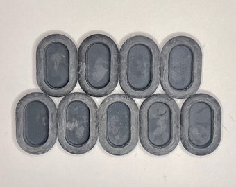 May include: Ten dark grey rubber grommets, oval in shape, are arranged on a white background. Each grommet has a raised border and a recessed centre. The grommets are evenly spaced and appear to be unused.