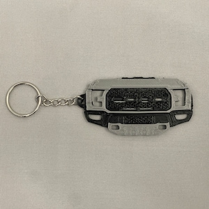 May include: A two-tone keychain featuring a detailed replica of a truck grille. The keychain is primarily gray and black, with a silver key ring and chain. The design includes the truck's front grill and bumper.