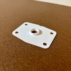 May include: A white, rectangular plastic component with a central circular opening and four evenly spaced holes at the corners. The object is set against a brown background, suggesting a product or part.