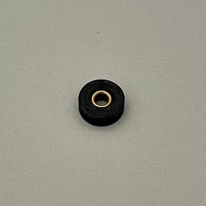 May include: A small, black, circular object with a gold-colored center hole. The object has a textured surface and appears to be made of a solid material. The item is set against a plain, light gray background.