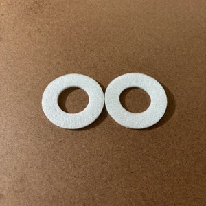 May include: Two white, circular felt pads with a hole in the center, arranged side by side. The pads have a textured surface and are set against a brown background.