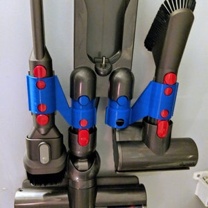 May include: A wall-mounted vacuum cleaner accessory holder with various attachments. The holder is gray with blue and red accents. The attachments include a brush head, a crevice tool, and a motorized cleaner head. The accessories are designed for a Dyson vacuum cleaner.