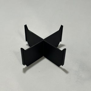 May include: A black, cross-shaped object with four upright supports. The object appears to be made of a solid material and is designed to hold something in place. The supports are rectangular and extend upwards from the cross.