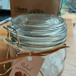 May include: A glass jar with a white label that says "Naked Sourdough Artisan Bread" with a brown bread illustration and the Instagram handle "@nakedsourd". The jar is tied with a brown ribbon and contains a white sourdough starter.