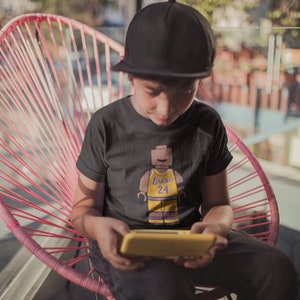 Peut inclure: Une jeune personne portant un t-shirt noir avec une figurine Lego d'un joueur de basket-ball portant un maillot jaune avec le numéro 24 et le texte "LeBron" dessus. La personne est assise sur une chaise rose et tient un appareil de jeu portable jaune.