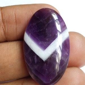 Puede incluir: Una piedra preciosa ovalada en cabuj&oacute;n de color p&uacute;rpura y blanco con un patr&oacute;n de chevron. La piedra preciosa est&aacute; pulida y tiene un acabado brillante.