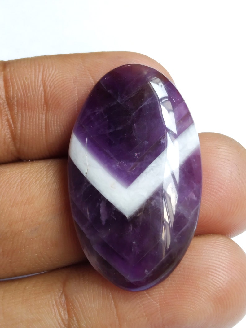 Puede incluir: Una piedra preciosa ovalada en cabuj&oacute;n de color p&uacute;rpura y blanco con un patr&oacute;n de chevron. La piedra preciosa est&aacute; pulida y tiene un acabado brillante.