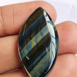May include: A blue and gold tiger's eye cabochon gemstone with a pointed oval shape. The gemstone has a chatoyant effect, meaning it shimmers and changes color depending on the angle of light.