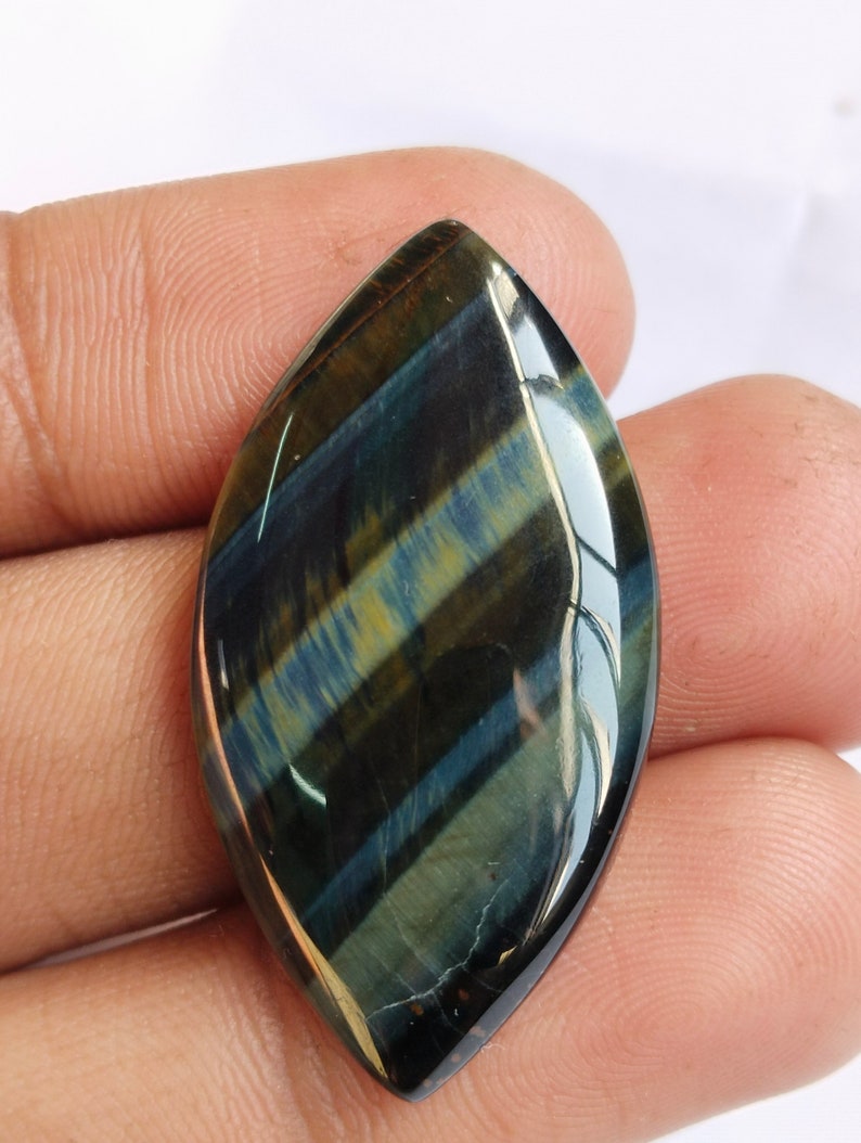 May include: A blue and gold tiger's eye cabochon gemstone with a pointed oval shape. The gemstone has a chatoyant effect, meaning it shimmers and changes color depending on the angle of light.