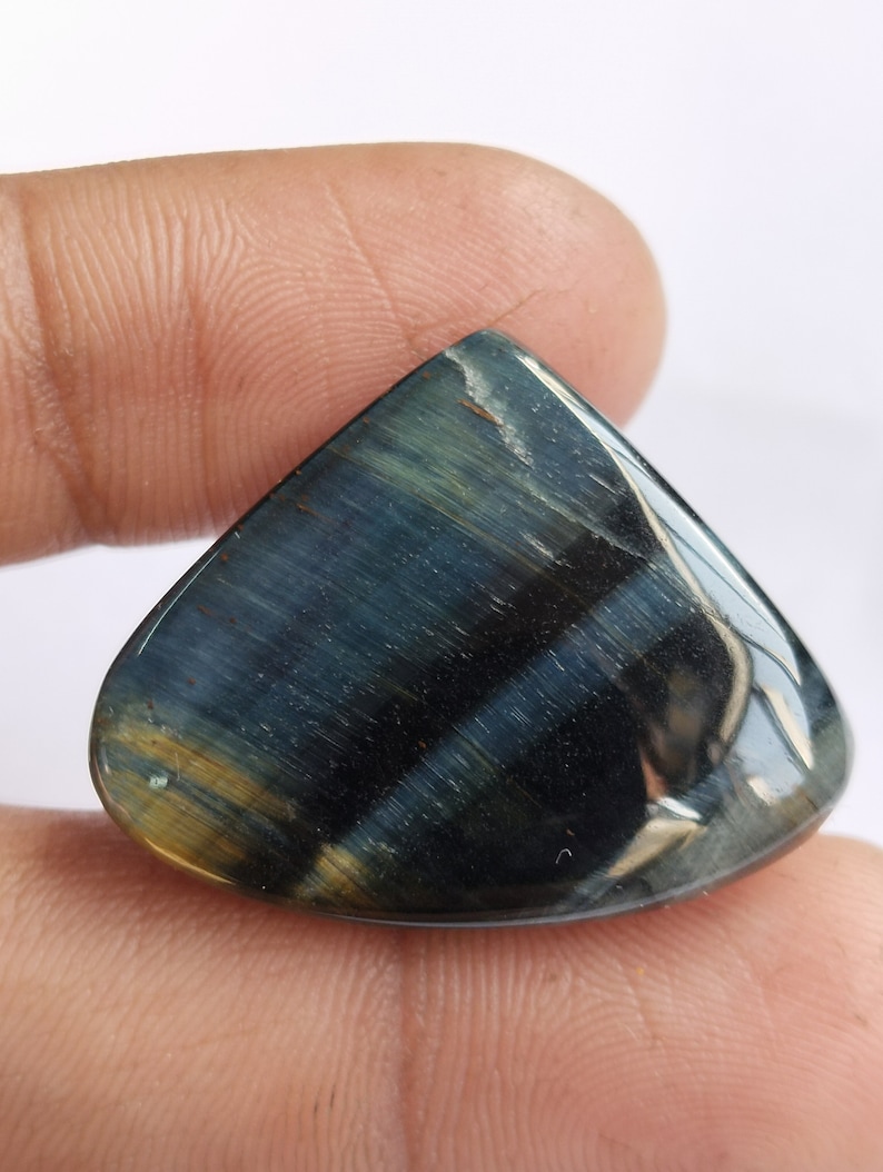 May include: A blue and gold tiger's eye gemstone cabochon with a teardrop shape. The stone has a reflective surface and a striated pattern.