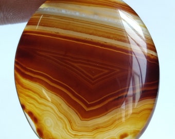 May include: A smooth, oval-shaped agate gemstone with a banded pattern of brown, orange, and white. The agate has a polished finish and is translucent.