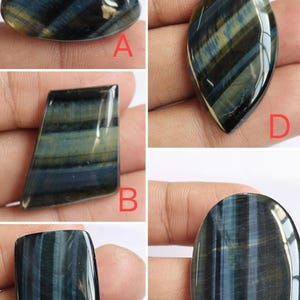 May include: Five different blue tiger's eye cabochons, each with a unique shape and pattern. The cabochons are displayed on a hand, with the letters A, B, C, D, and E labeling each stone.