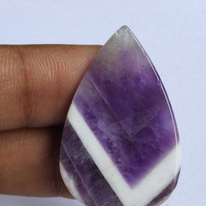 Puede incluir: Una piedra preciosa de amatista cabuj&oacute;n de forma de l&aacute;grima, de color p&uacute;rpura y blanco. La piedra preciosa tiene un patr&oacute;n de chevron de blanco y p&uacute;rpura.