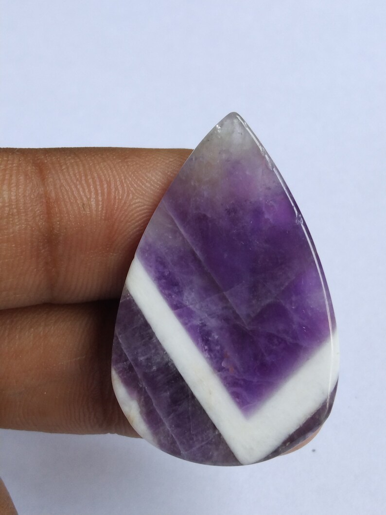 Puede incluir: Una piedra preciosa de amatista cabuj&oacute;n de forma de l&aacute;grima, de color p&uacute;rpura y blanco. La piedra preciosa tiene un patr&oacute;n de chevron de blanco y p&uacute;rpura.