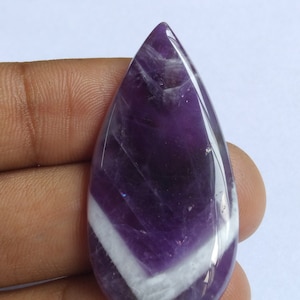 Puede incluir: Un cabuj&oacute;n de piedra preciosa amatista p&uacute;rpura y blanca con forma de l&aacute;grima. La piedra preciosa tiene una superficie lisa y pulida y una banda blanca que atraviesa el centro.