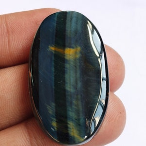 May include: Oval-shaped blue tiger's eye gemstone cabochon with a golden sheen. The stone has a smooth, polished surface and is approximately 2 inches long.