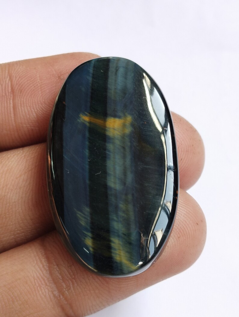 May include: Oval-shaped blue tiger's eye gemstone cabochon with a golden sheen. The stone has a smooth, polished surface and is approximately 2 inches long.