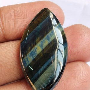 May include: A blue and gold tiger's eye cabochon gemstone with a teardrop shape. The gemstone has a smooth, polished surface and a unique, iridescent sheen.