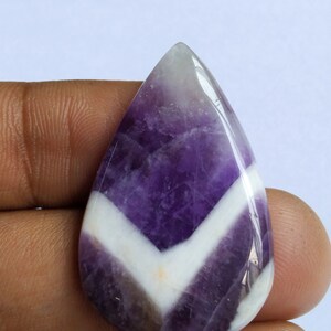 Puede incluir: Una piedra preciosa de amatista cabuj&oacute;n de color p&uacute;rpura y blanco con un patr&oacute;n de chevron. La piedra preciosa tiene forma de l&aacute;grima y una superficie lisa y pulida.