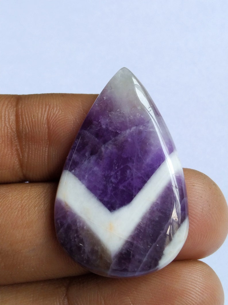 Puede incluir: Una piedra preciosa de amatista cabuj&oacute;n de color p&uacute;rpura y blanco con un patr&oacute;n de chevron. La piedra preciosa tiene forma de l&aacute;grima y una superficie lisa y pulida.