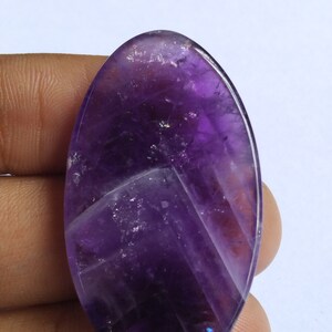 Puede incluir: Una gran piedra preciosa de amatista de forma ovalada, de color morado oscuro y tallada a facetas. La piedra tiene un aspecto natural, nublado, con inclusiones visibles.