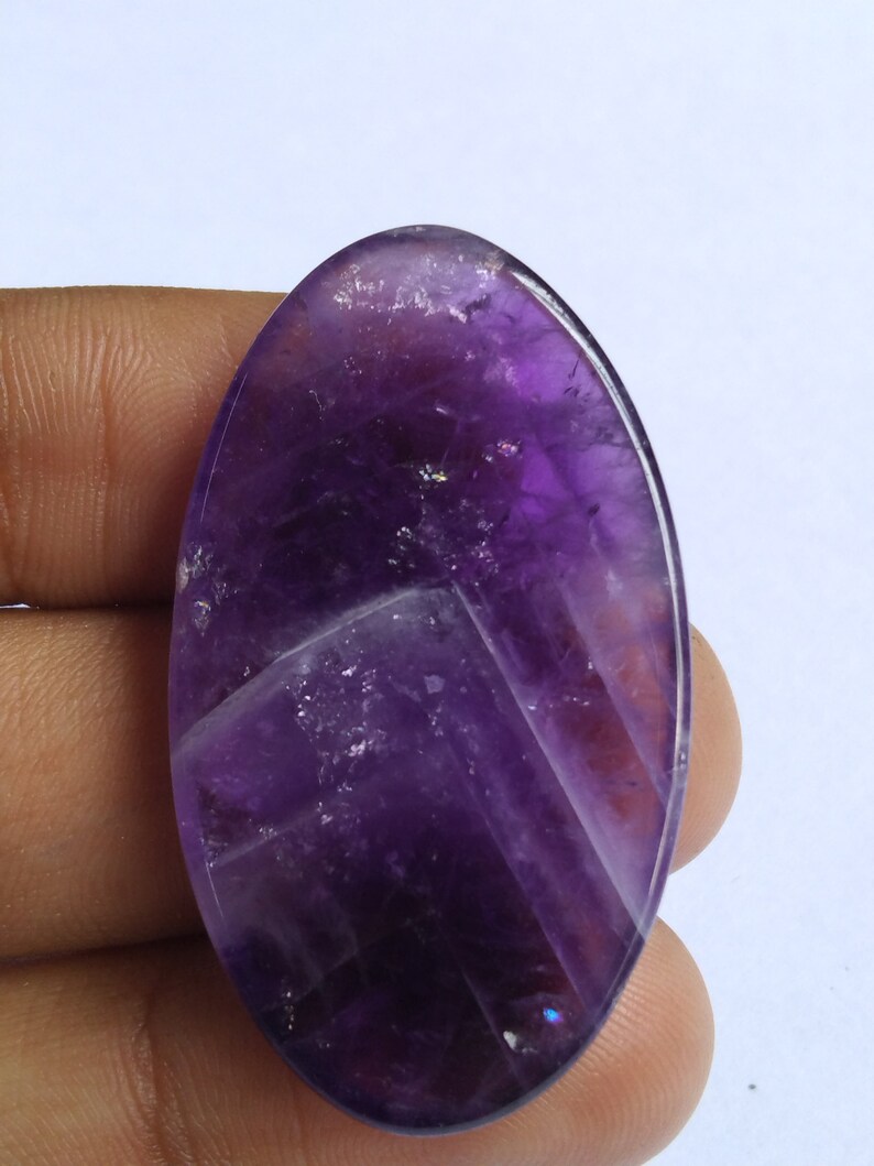 Puede incluir: Una gran piedra preciosa de amatista de forma ovalada, de color morado oscuro y tallada a facetas. La piedra tiene un aspecto natural, nublado, con inclusiones visibles.