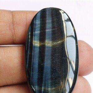May include: A blue and gold oval-shaped tiger's eye gemstone cabochon. The stone has a chatoyant effect, meaning it appears to have a shimmering, cat's eye-like effect.