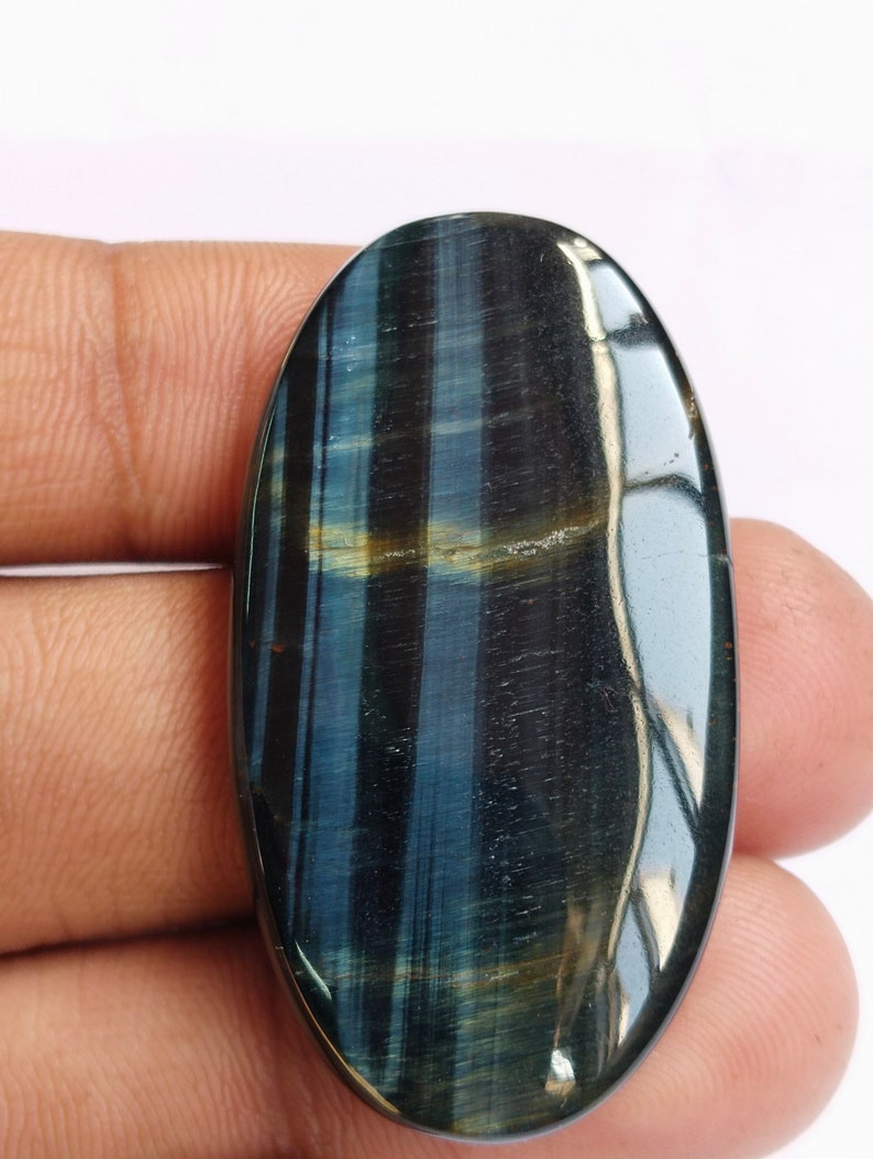 May include: A blue and gold oval-shaped tiger's eye gemstone cabochon. The stone has a chatoyant effect, meaning it appears to have a shimmering, cat's eye-like effect.