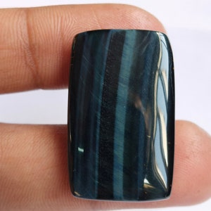 May include: A dark blue and black tiger's eye cabochon gemstone with a rectangular shape. The stone has a chatoyant effect, meaning it appears to have a shimmering or cat's eye effect.