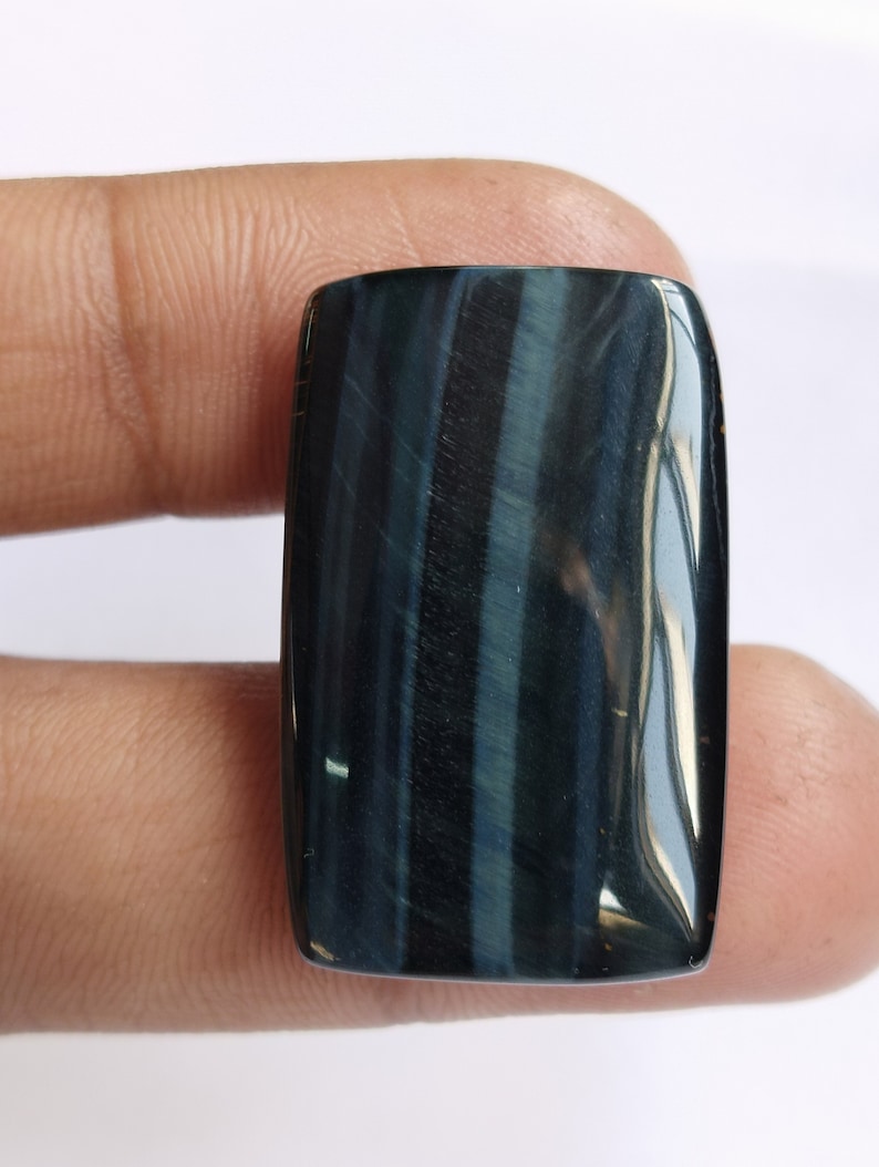 May include: A dark blue and black tiger's eye cabochon gemstone with a rectangular shape. The stone has a chatoyant effect, meaning it appears to have a shimmering or cat's eye effect.