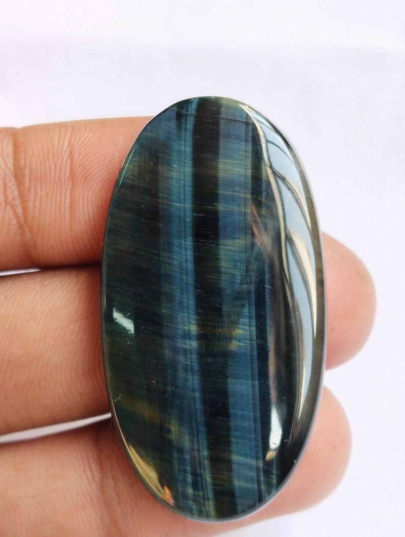 May include: A blue and green oval-shaped gemstone with a tiger's eye pattern. The gemstone is smooth and polished.