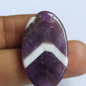 Puede incluir: Una piedra preciosa de amatista de forma ovalada con un patr&oacute;n de chevron en color morado y blanco.