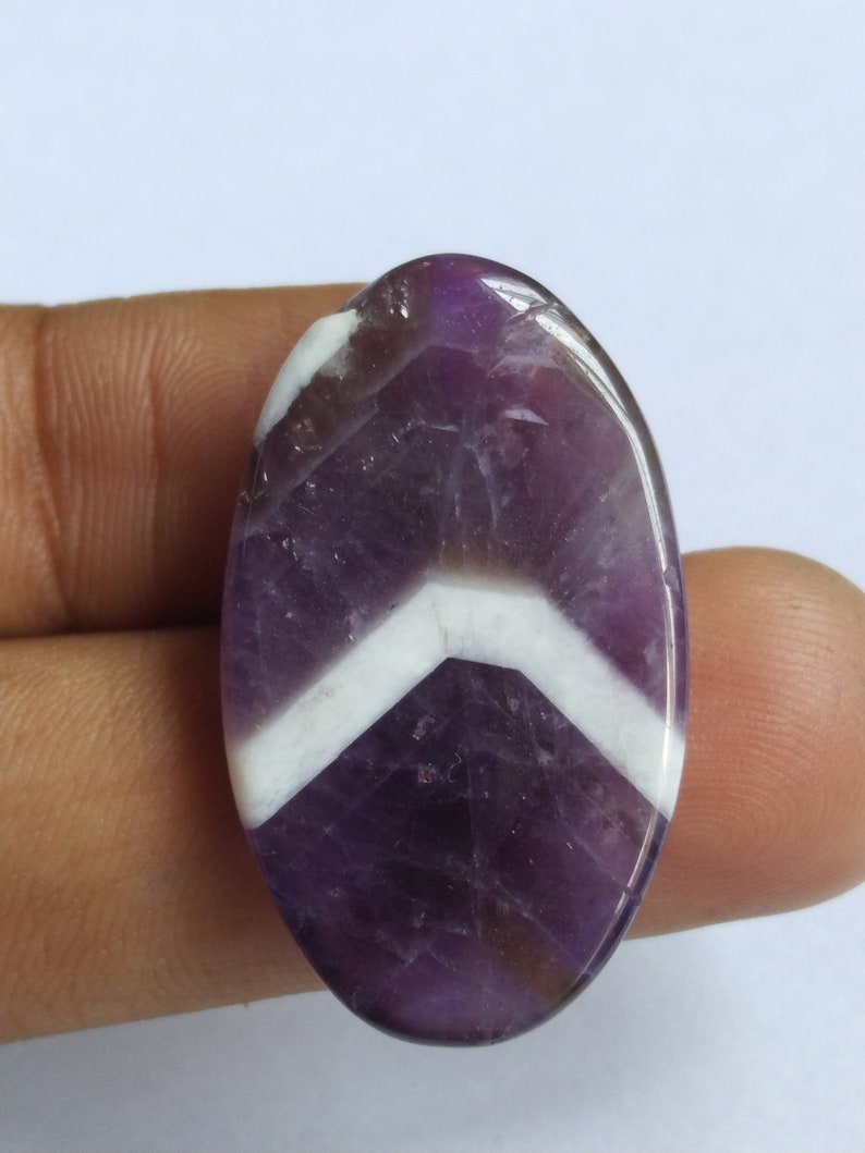 Puede incluir: Una piedra preciosa de amatista de forma ovalada con un patr&oacute;n de chevron en color morado y blanco.