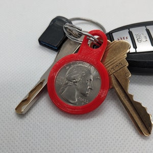 May include: A red keychain with a 2022 quarter dollar coin embedded in the center. The coin is surrounded by a thick red plastic ring. The keychain is attached to a set of keys.