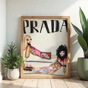 以下が含まれることがあります： 大きな黒い筆致の文字でPRADAと書かれた額入りアートプリント。その下には2人の人物の水彩画イラストが描かれています。一人はブロンドの髪にピンクのトップスを着ており、もう一人は黒髪で柄物の衣装を身に着けています。