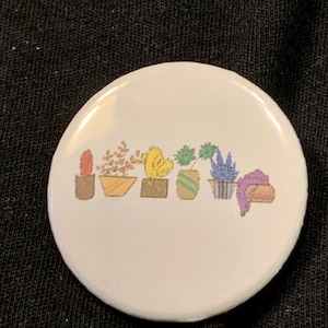 Puede incluir: Un botón blanco con una línea de siete plantas en maceta de diferentes colores y estilos. Las plantas están en fila de izquierda a derecha, y todas están en macetas diferentes. Las macetas son todas de diferentes colores y estilos.