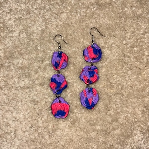 May include: A pair of dangle earrings featuring three stacked, abstract, circular shapes. Each shape is a swirl of purple, pink, and blue. The earrings have silver-colored ear wires.
