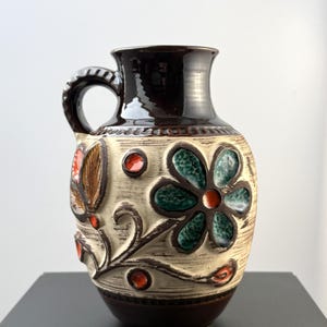 Könnte beinhalten: Ein Keramikkrug mit dunkelbraun glasiertem Hals und Boden. Der cremefarbene Körper hat eine texturierte Oberfläche mit horizontalen Linien und geprägten Blumenmotiven in Dunkelgrün und leuchtendem Orange sowie einen geschwungenen Henkel.