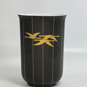 Könnte beinhalten: Schwarze Keramikvase mit vertikalen Goldstreifen und einem goldenen Design fliegender Gänse. Die Vase hat einen weißen Innenraum und einen abgerundeten Boden. Das Design ist schlicht und elegant.