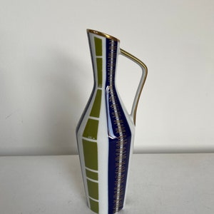 May include: A white ceramic vase with a geometric design in shades of green and blue. The vase has a gold trim and a handle.