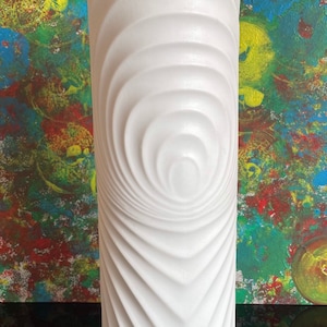 May include: A tall, white ceramic vase with a textured, swirling design. The vase has a cylindrical shape and a geometric base. The background features an abstract painting with vibrant colors. The vase is approximately 12 inches tall.