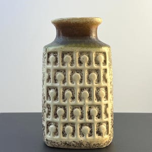 May include: A rectangular ceramic vase with a cream-colored body featuring a raised grid pattern of circles and vertical lines. The neck of the vase transitions into a dark brown and green glaze, while the base shows a speckled brown texture.