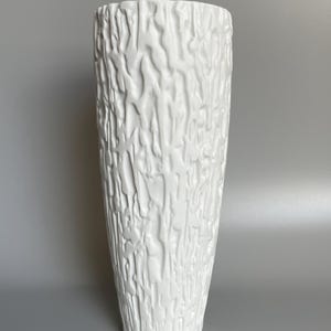 Könnte beinhalten: Eine weiße Keramikvase mit einem strukturierten, baumrindenartigen Design. Die Vase ist hoch und zylindrisch.