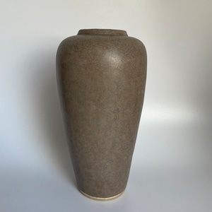 Könnte beinhalten: Eine hohe, strukturierte Vase in einem gedämpften Braunton. Die Vase hat eine leicht konische Form mit abgerundeter Oberseite und einem goldfarbenen Band am Boden. Die Oberfläche hat ein gesprenkeltes Aussehen.