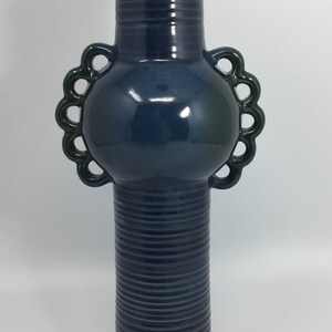 Könnte beinhalten: Eine hohe, dunkelblaue Keramikvase mit glänzendem Finish. Die Vase hat einen zylindrischen Körper mit horizontalen Rillen, einen abgerundeten Mittelteil mit dekorativen Griffen und einen schmaleren, geriffelten Hals. Die Griffe sind in einem dunkleren Grünton gehalten.